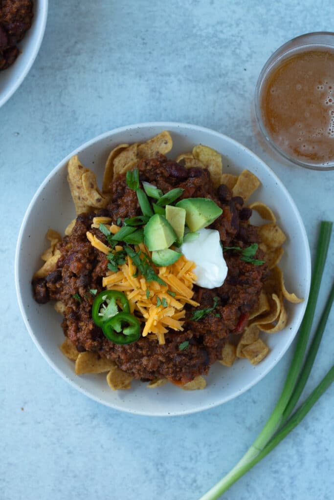 Thick hearty chili on top of corn chips with toppings and a glass of beer on side