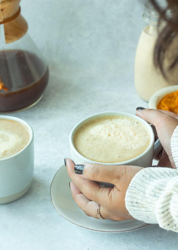 hands picking up coffee mug with coffee pot and pumpkin in back