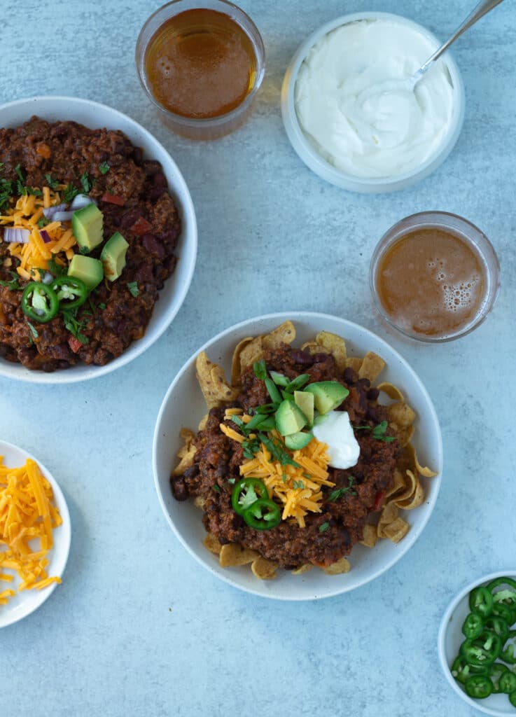 Two bowls of chili with beer and toppings on side.