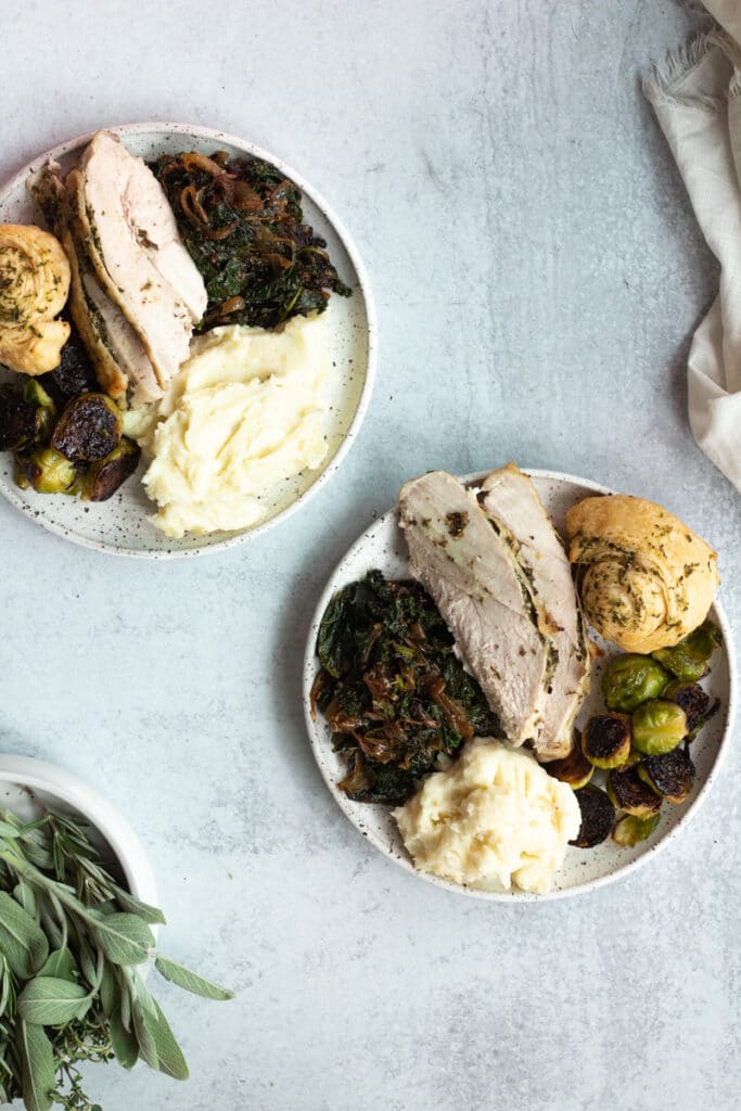 two plates with thanksgiving food including turkey, mashed potatoes, sprouts and bread with a small bowl of herbs off to the side