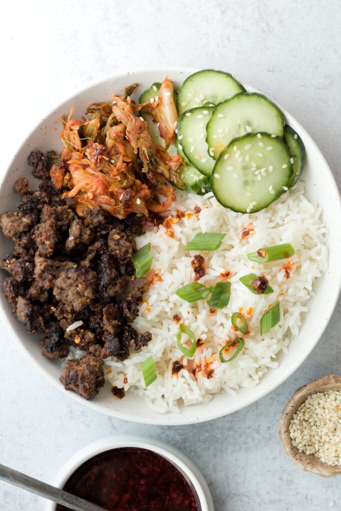 Bowl with ground beef bulgogi, rice, cucumber slices and sesame seeds with chili oil on side