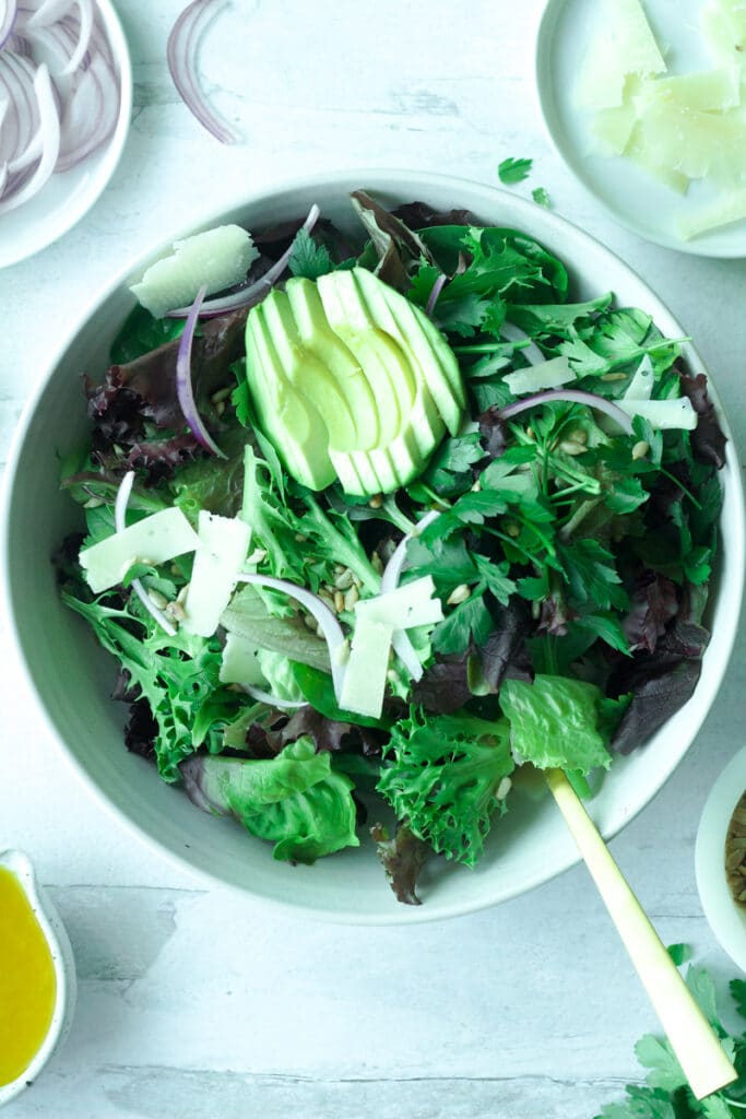 Big bowl of salad greens topped with sliced avocado and shaved parmesan cheese with gold fork on side