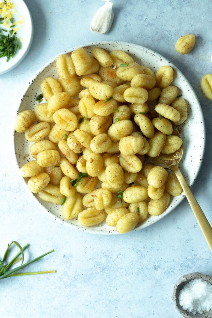 roasted gnocchi with lemon garlic brown butter on plate surrounded by chives, a small bowl of flaky salt and garlic cloves