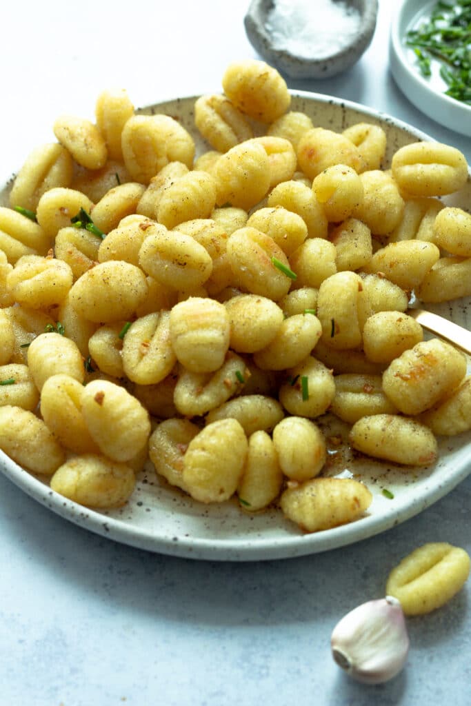 large plate of roasted gnocchi tossed with butter, garlic and lemon with small bowls of chopped chives and flaky salt in background