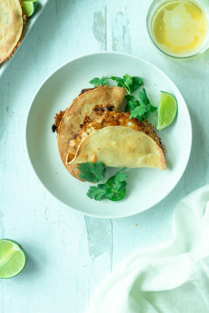 Two baked chicken tacos on round plate with lime wedge and cilantro leaves next to white napkin and beer glass