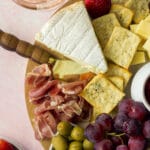 Wedge of brie of cheese board next to olives, grapes and prosciutto with glass of rose
