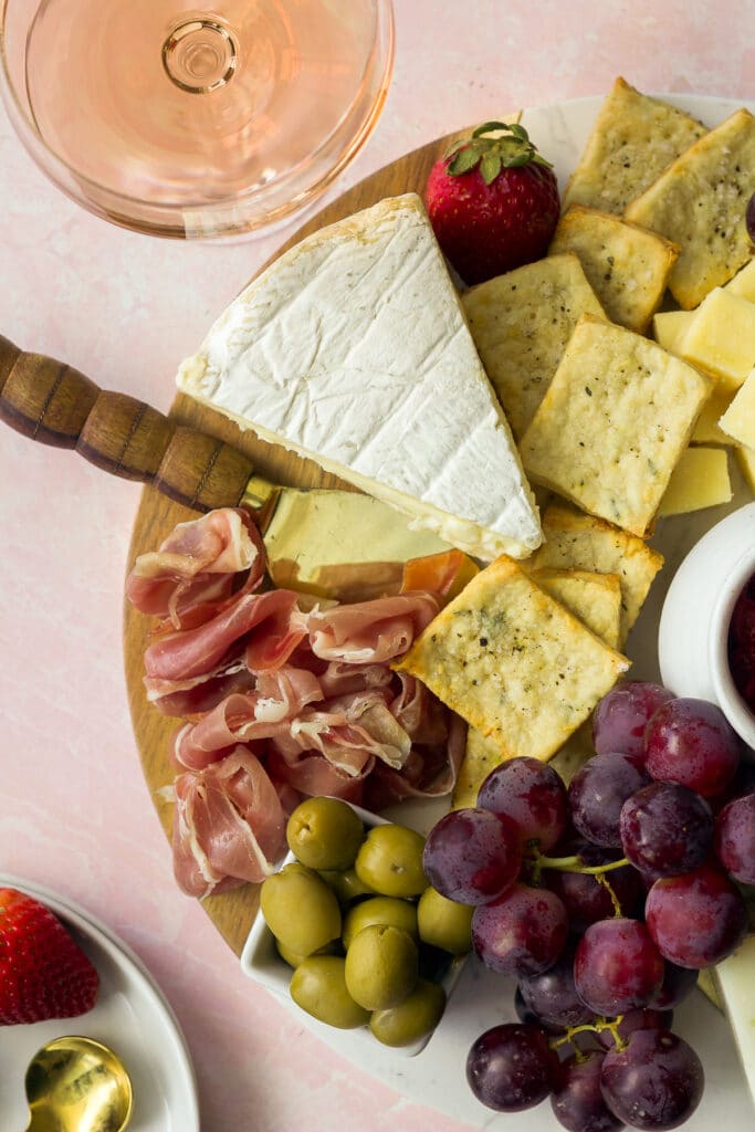 Wedge of brie of cheese board next to olives, grapes and prosciutto with glass of rose