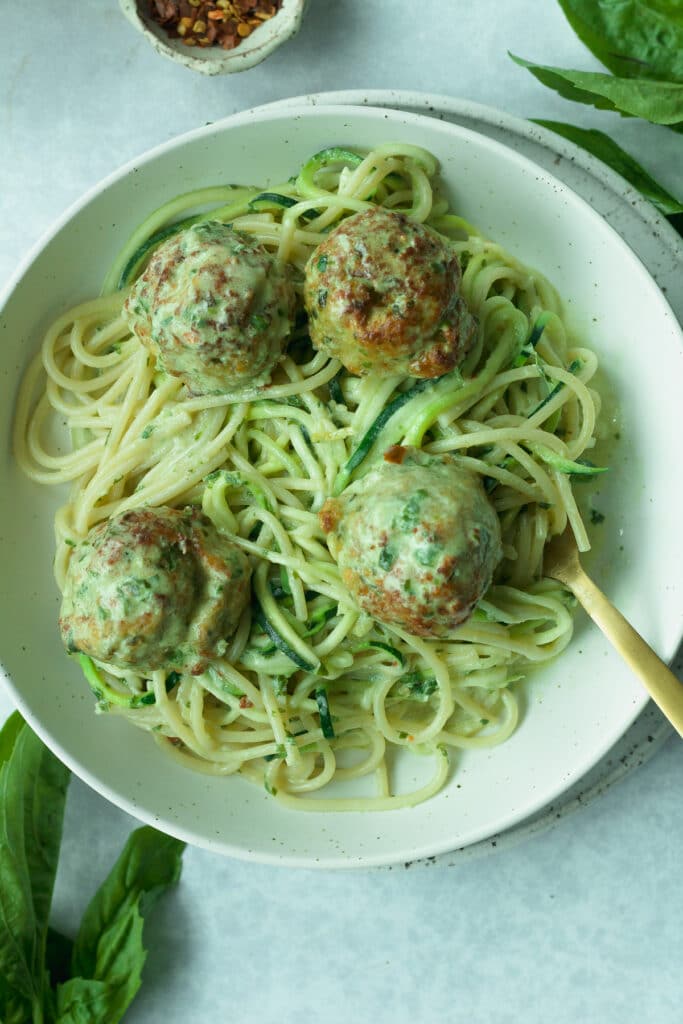 Bowl of pasta with chicken meatballs with fork in bowl and basil and red pepper flakes scattered around.