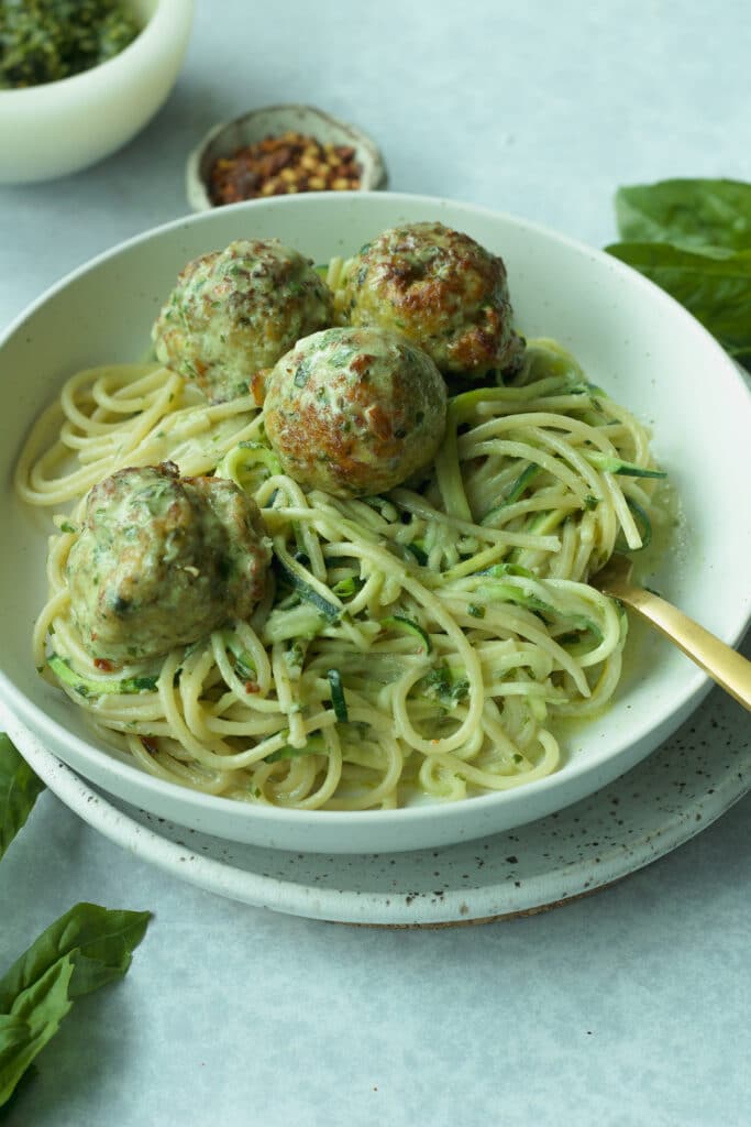 45 degree angle of creamy pesto chicken meatball pasta with bowl of red pepper flakes and pesto in background