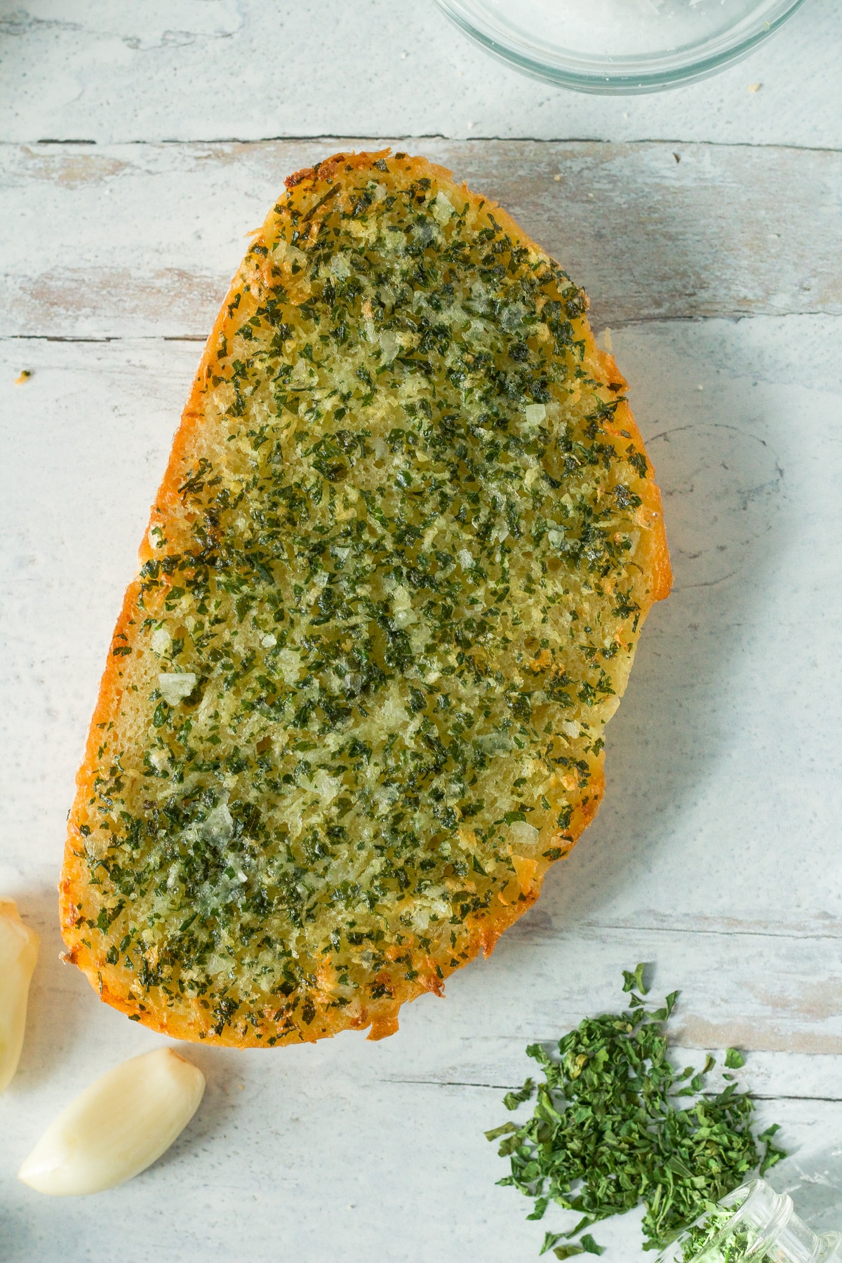 Garlic bread on board with garlic cloves and dried parsley around it.