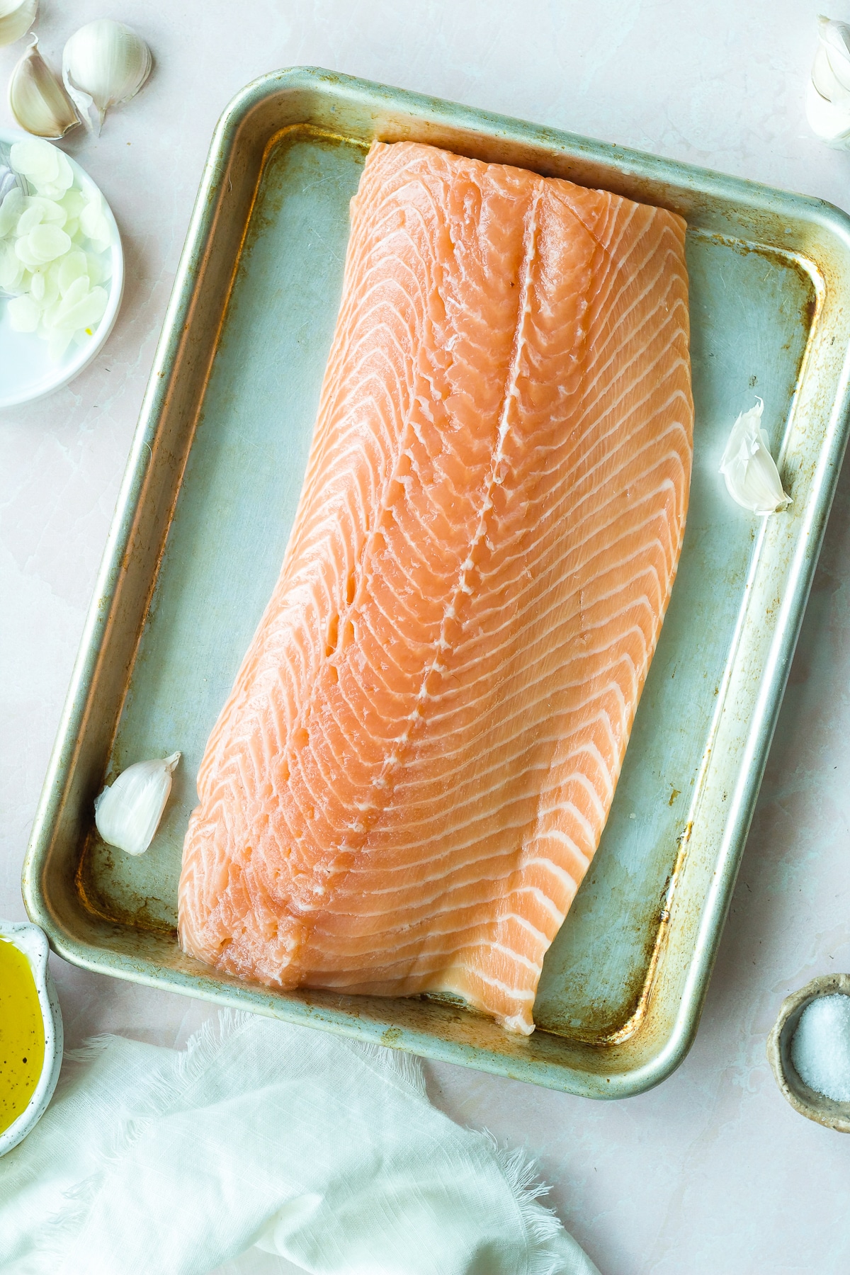 Raw salmon on baking sheet with garlic and small bowl of olive oil on side.