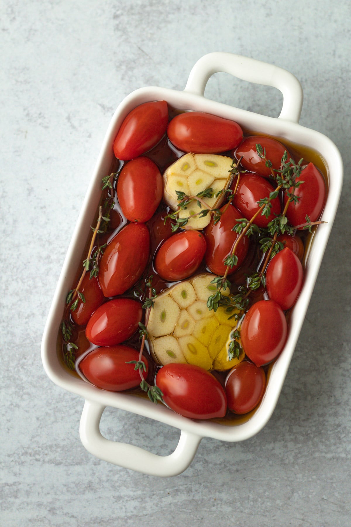 Cherry tomatoes, garlic and thyme covered in olive oil in dish.