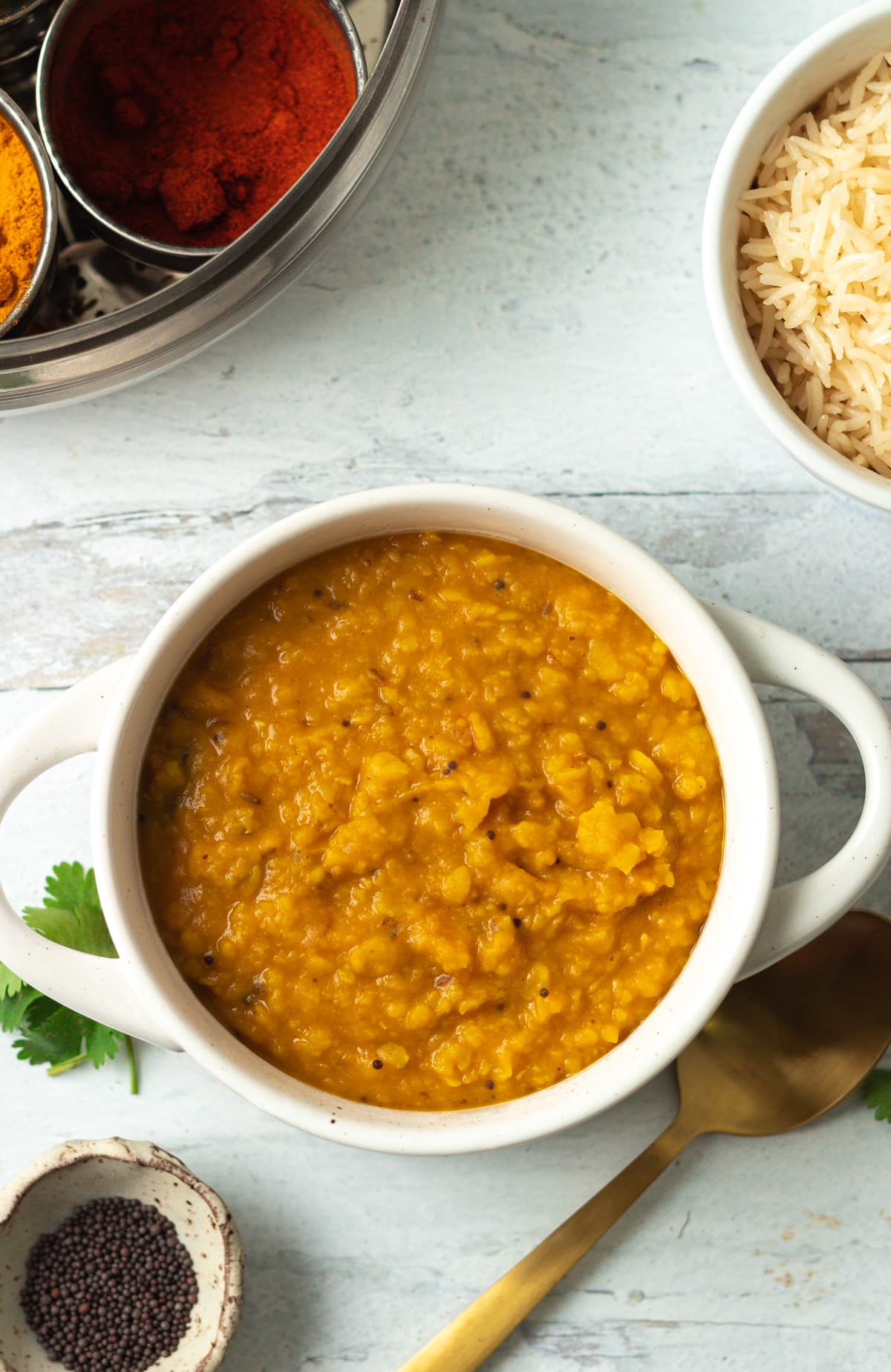 Dal in small bowl with bowl of rice, spices and spoon on side.